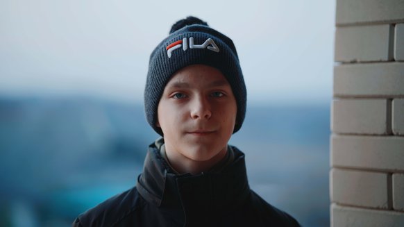 Ihor, a teenage boy from Ukraine, stands outside a flat in Moldova, slightly smiling, wearing a hat